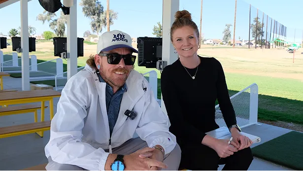 Smiling man in a white jacket and woman in black sitting together outdoors at a golf driving range.