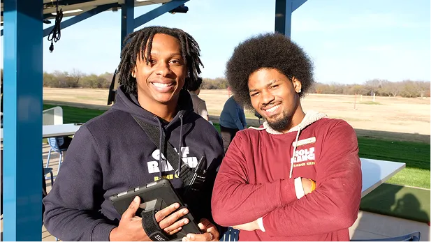 Two smiling men outdoors under shelter, one holding a tablet, both wearing hoodies.