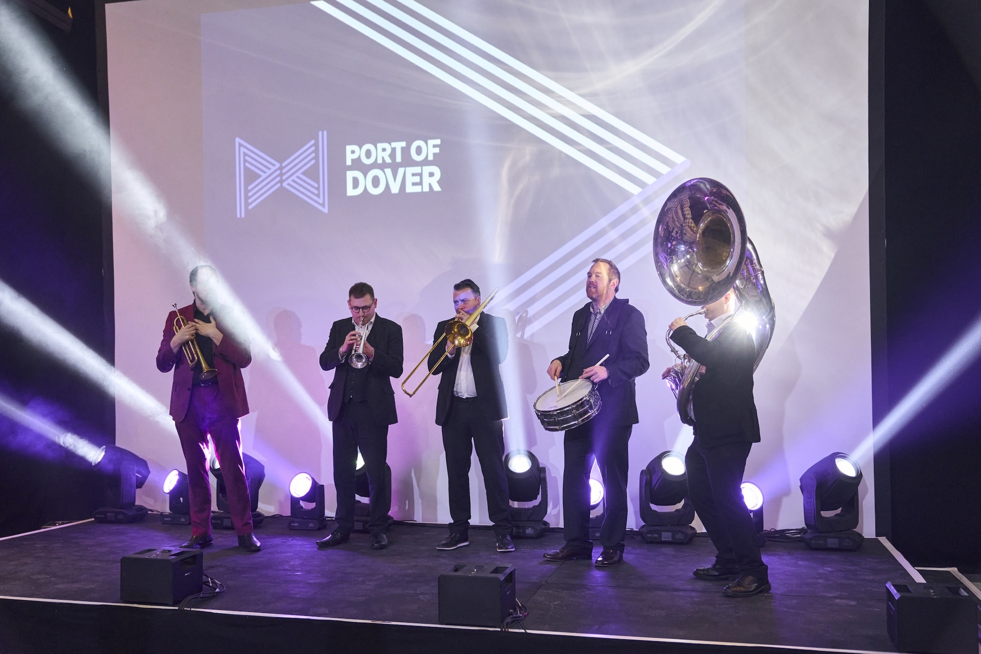 A five-piece brass band performing on the main stage at the Port of Dover Sea Change launch, with the Port of Dover logo projected behind them.