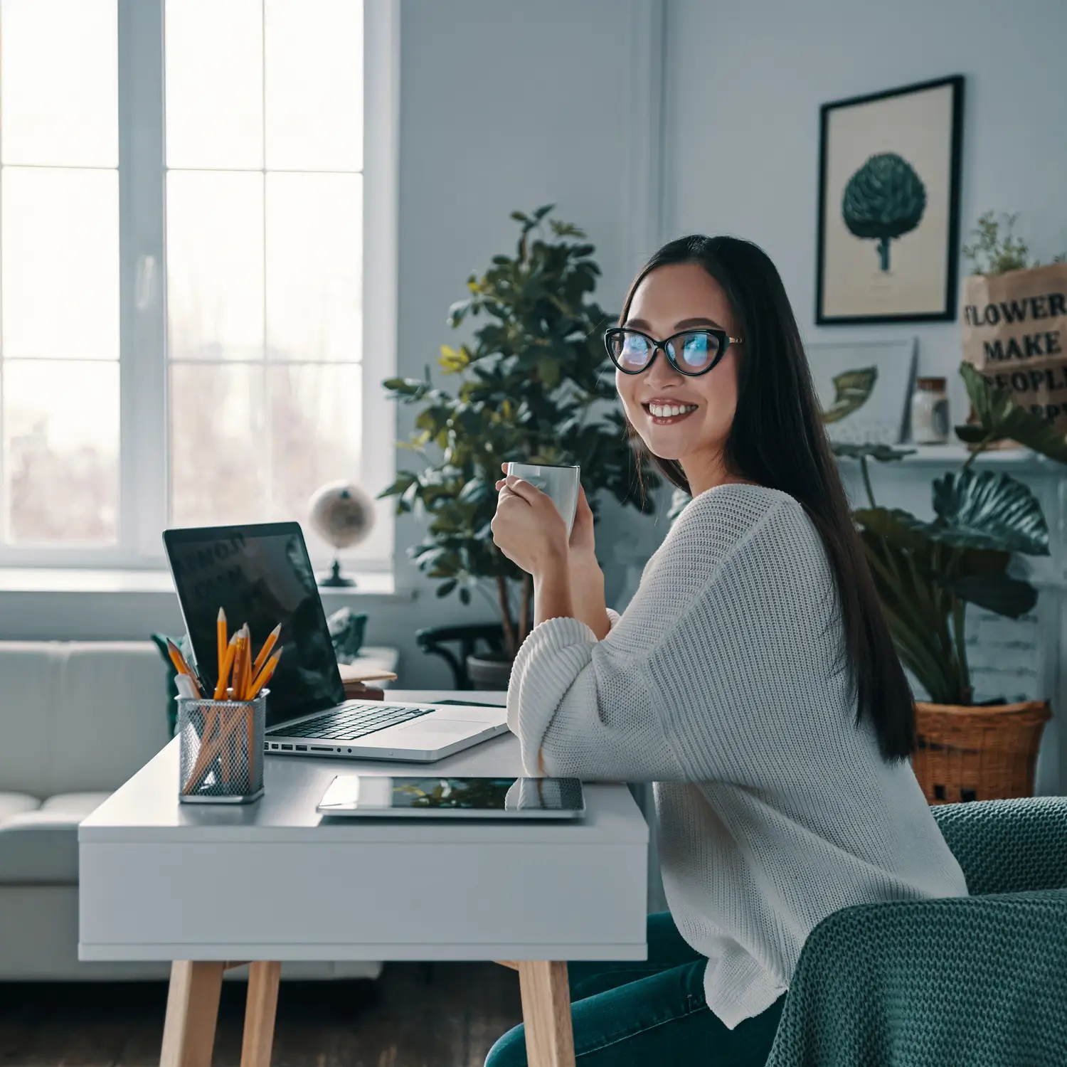 Smiling woman working from home, holding a coffee cup and sitting at her desk with a laptop and tablet.