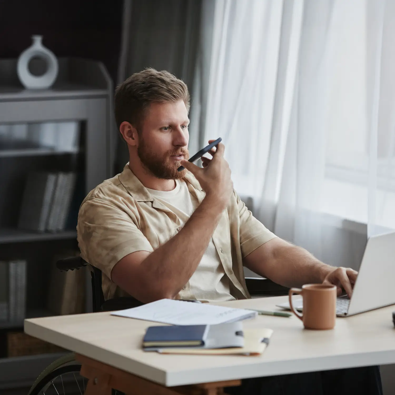 Man working remotely, using voice message on smartphone while analyzing notes and working on a laptop.