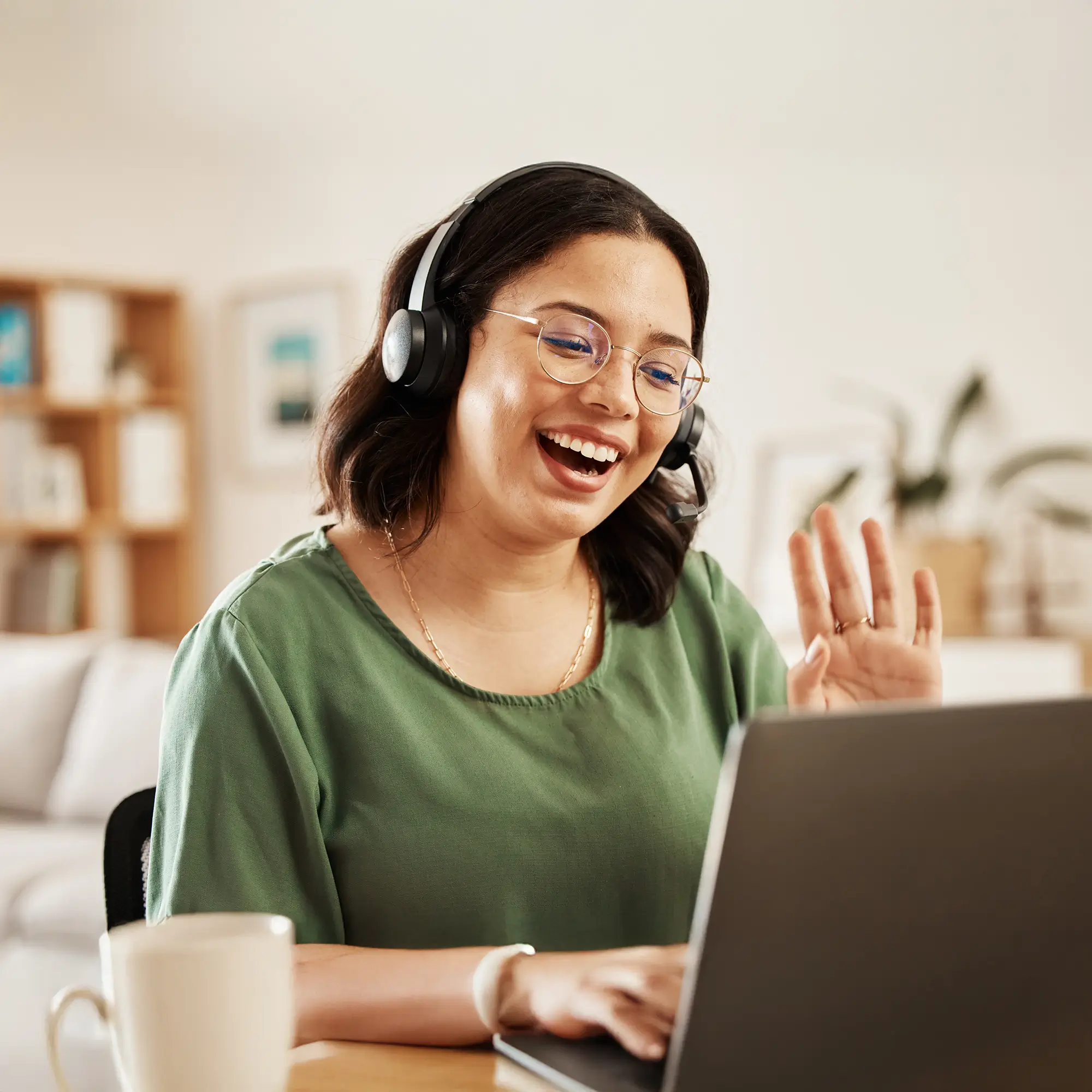 Smiling offshore customer service representative wearing a headset, waving during a video call from home office.
