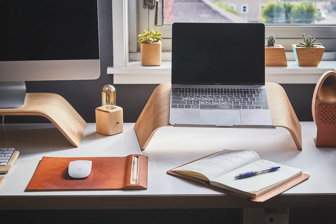 A working desk with a laptop and iMac. An notebook lies open next to a mouse.