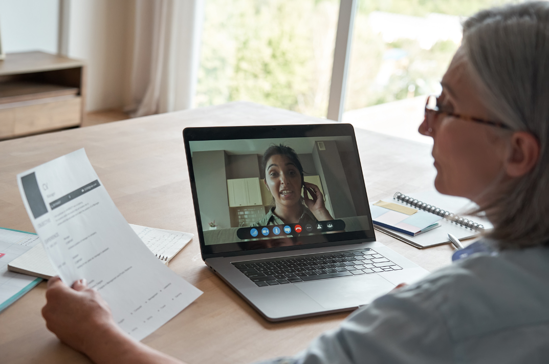 A female interviewer is at her desk, her laptop open as she glances to the left at a printed resume. A candidate is speaking through the video call.