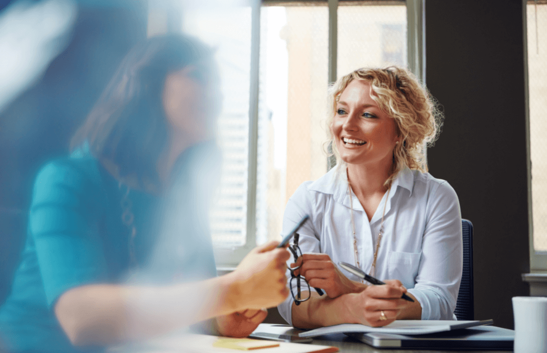 Two women seated side by side are engaged in conversation. The woman on the left is blurred, while the woman on the right smiles widely, watching her companion closely.