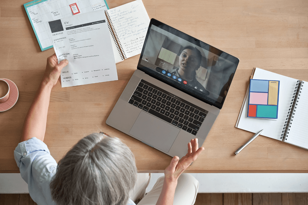 A woman, holding a printed CV, is interviewing an applicant online for a remote job.