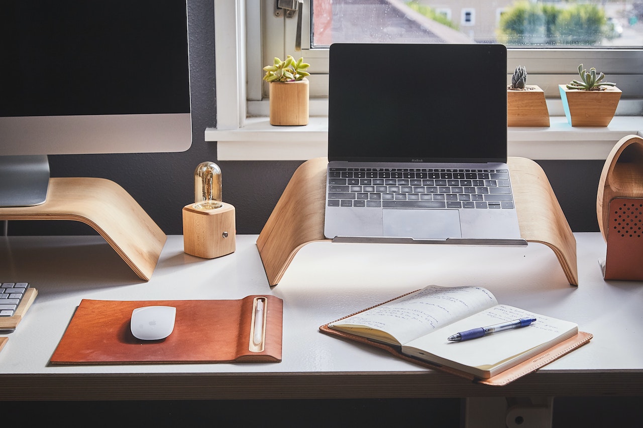 A working desk with a laptop and iMac. An notebook lies open next to a mouse.