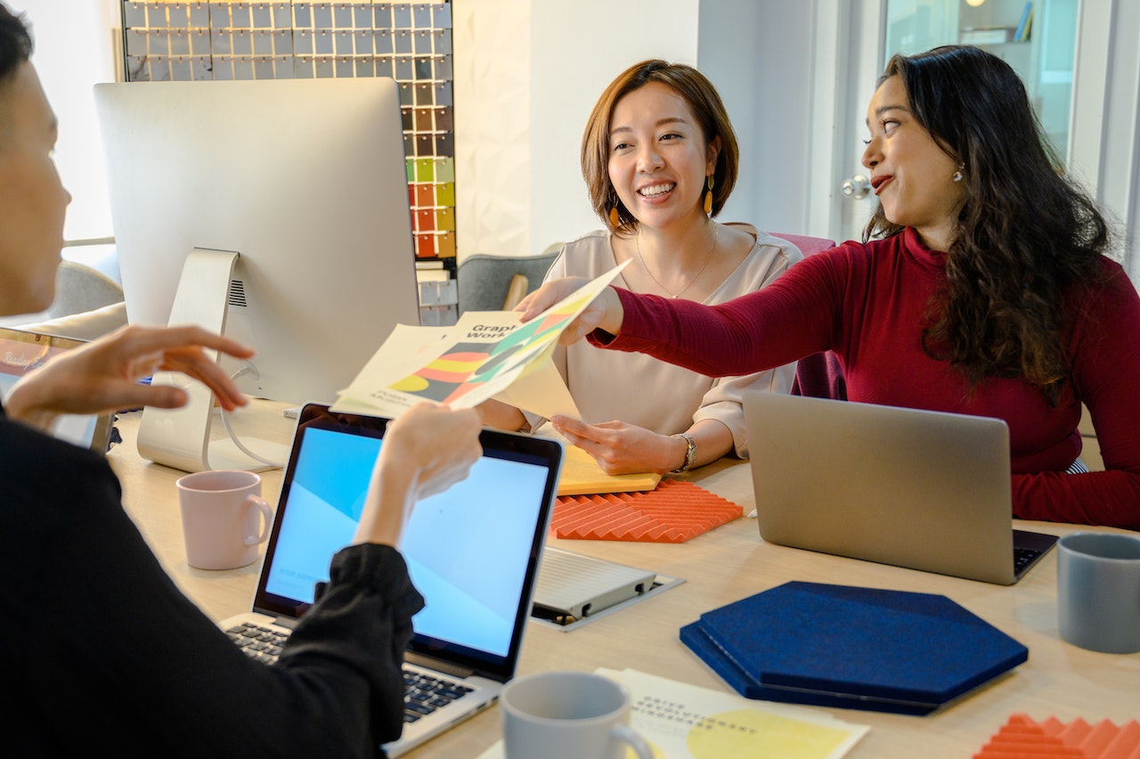 Three women working togehter in a design office. One of them passes a printed document to the person in front of her