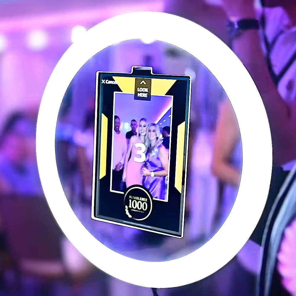 A digital photo booth screen surrounded by a ring light showing a group of people posing and a countdown timer at 3.