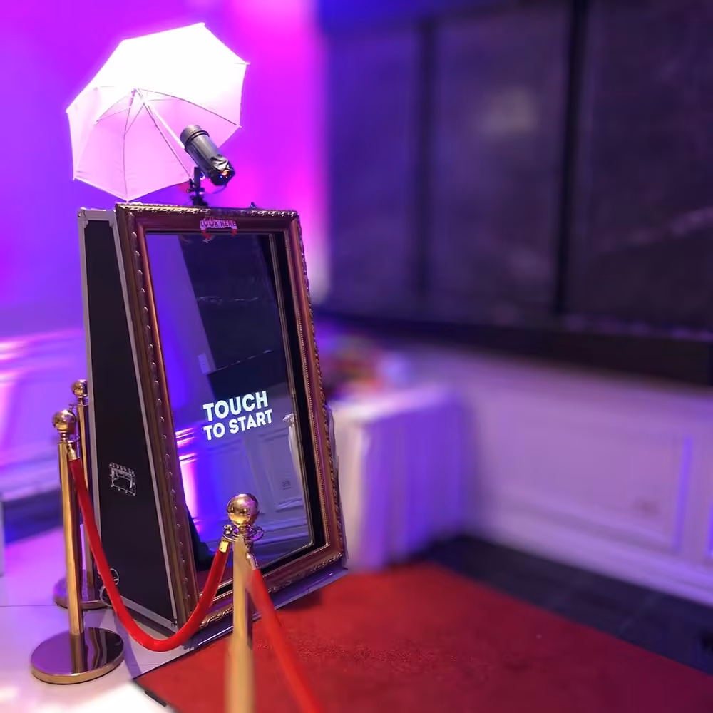 Interactive photo booth mirror with 'Touch to Start' text, a white umbrella light, and red velvet ropes on a red carpet.