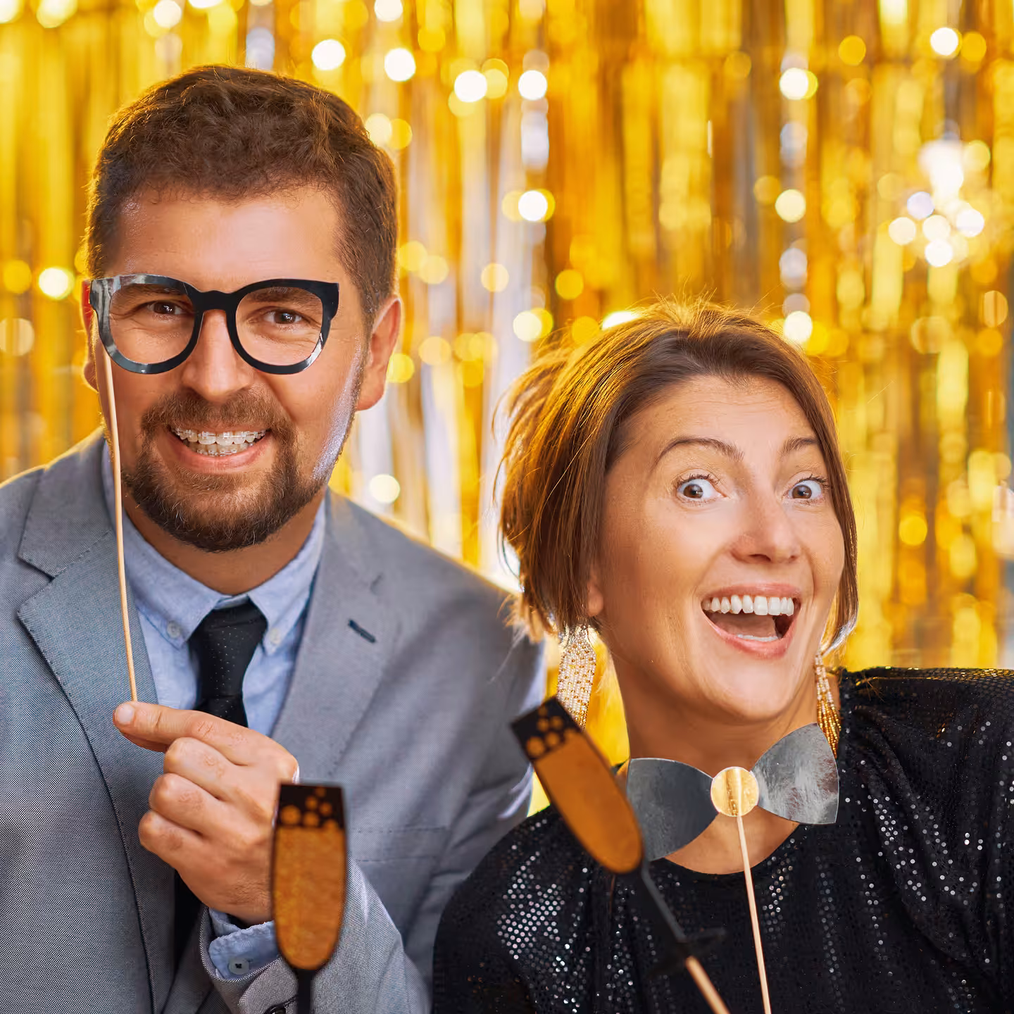 Smiling man and woman holding party props against a gold glittery background.