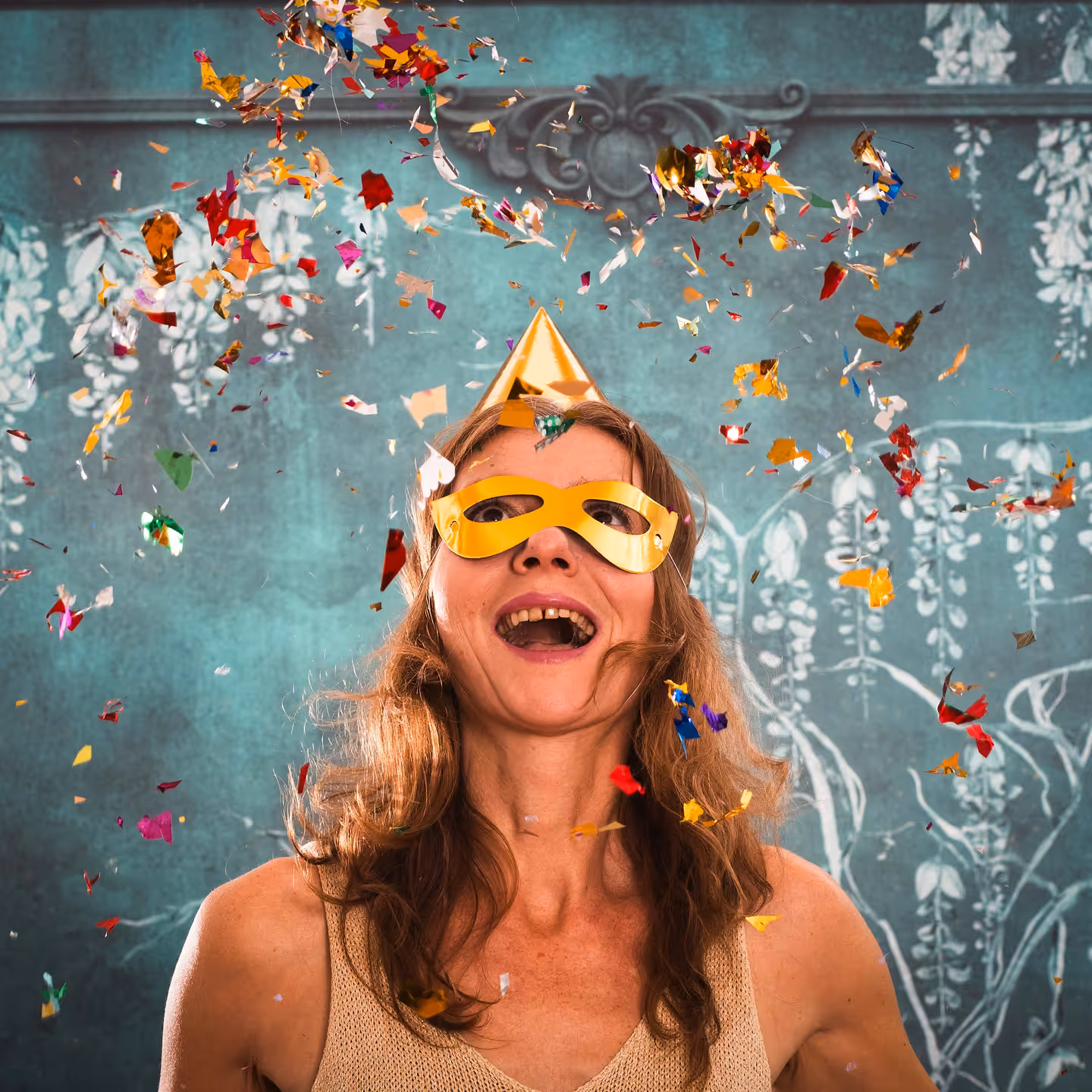 Woman wearing a golden party hat and mask smiling with colorful confetti falling around her against a teal decorative wall.