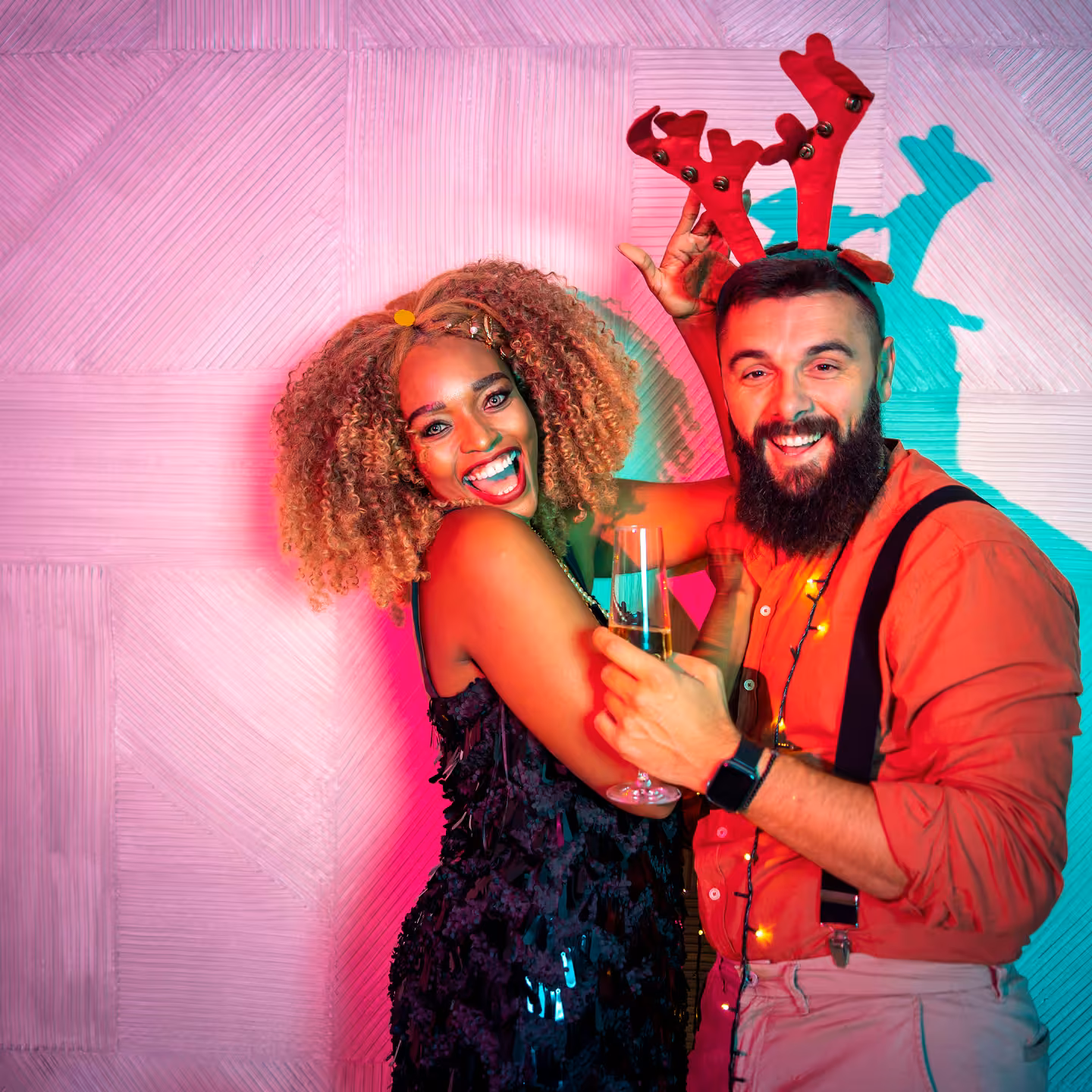 Smiling couple celebrating, woman in black dress and man with reindeer antlers holding champagne glass.