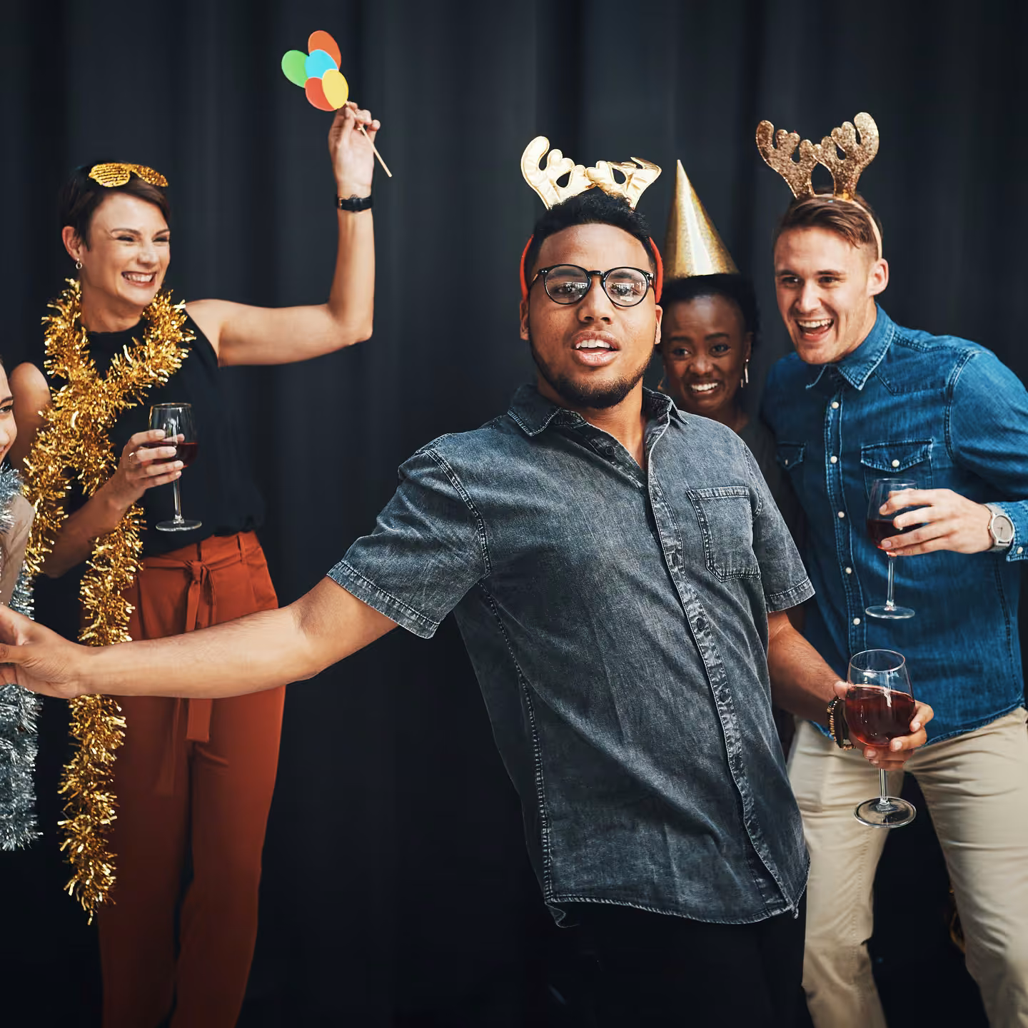 Group of friends wearing party hats and accessories, holding wine glasses and dancing at a celebration.