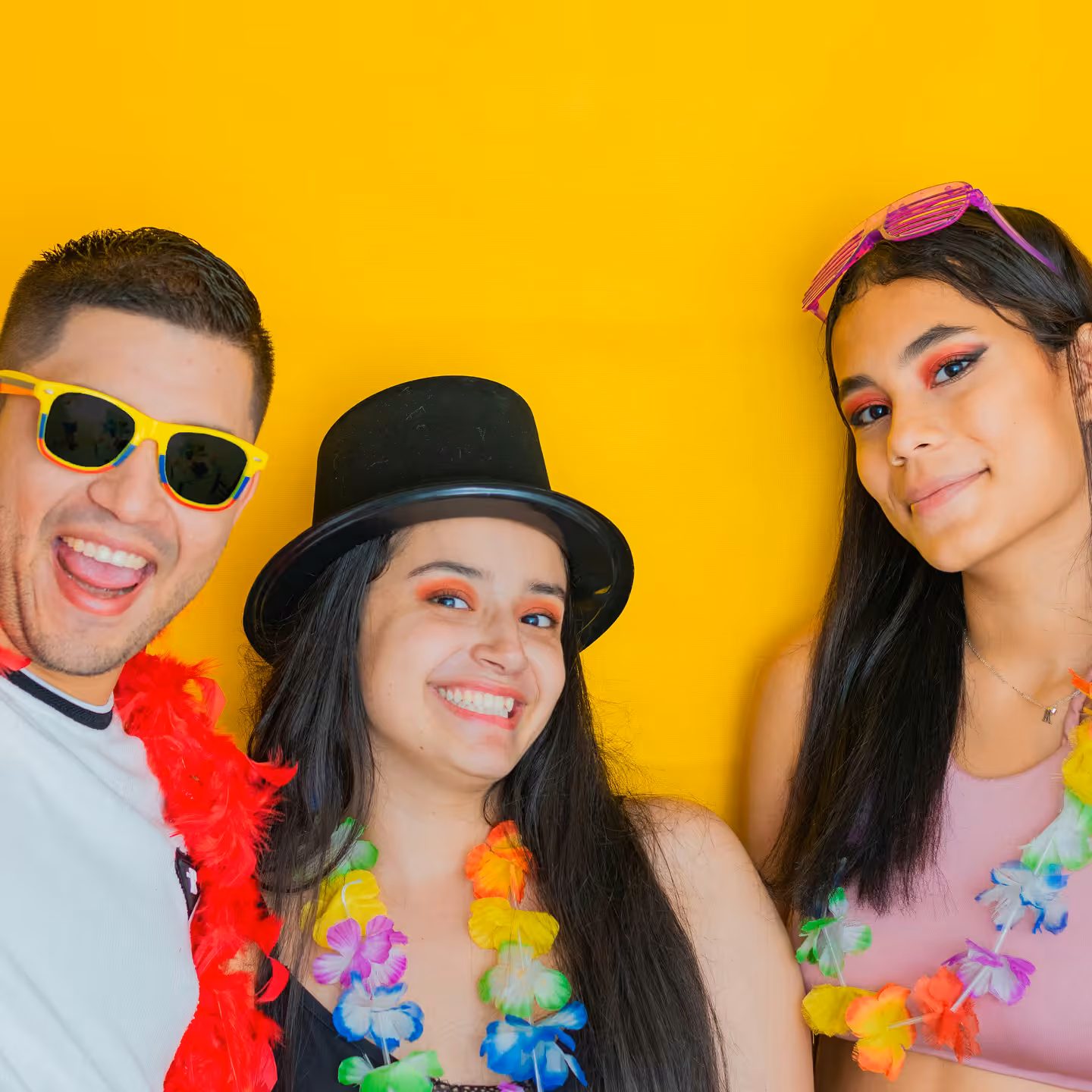 Three friends in leis, sunglasses and a top hat posing against a yellow backdrop