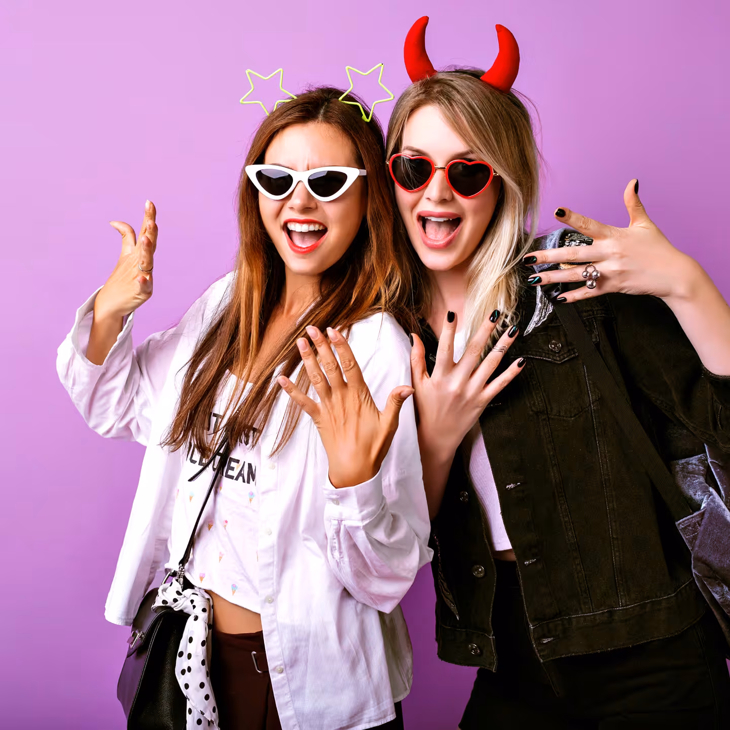 Two young women wearing novelty headbands and sunglasses show their manicured nails excitedly against a purple background.