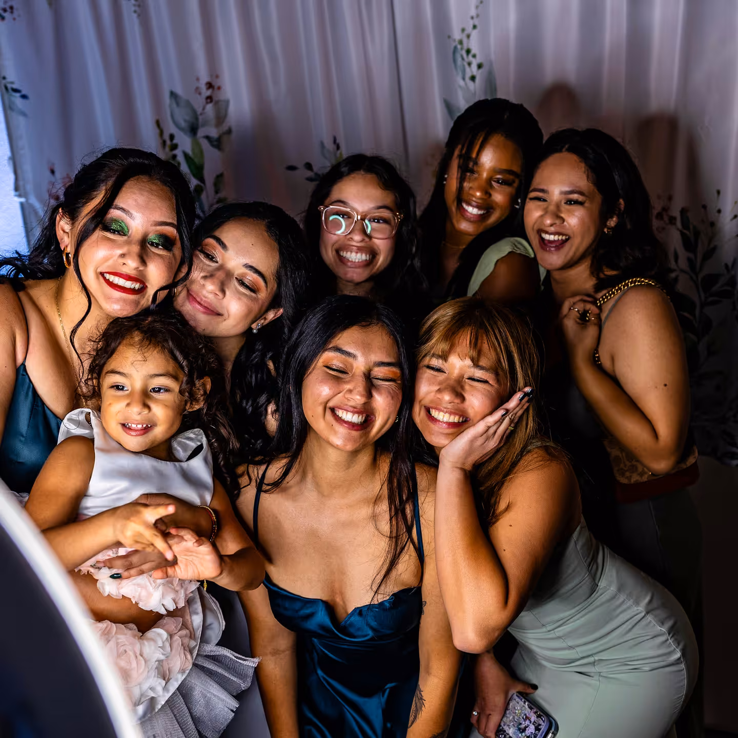 Group of eight women and a young girl smiling and posing closely together in front of a floral curtain backdrop.
