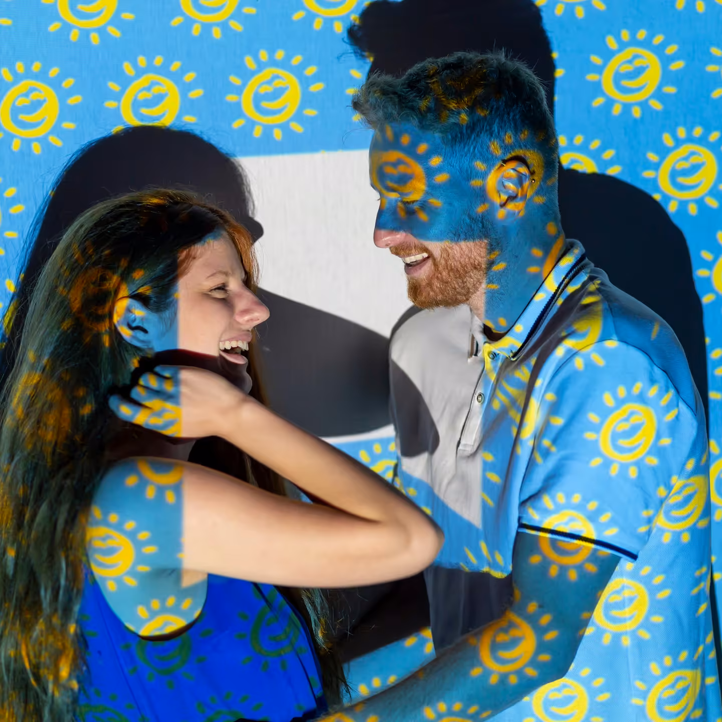Smiling couple looking at each other with yellow sun patterns projected onto them against a blue background.