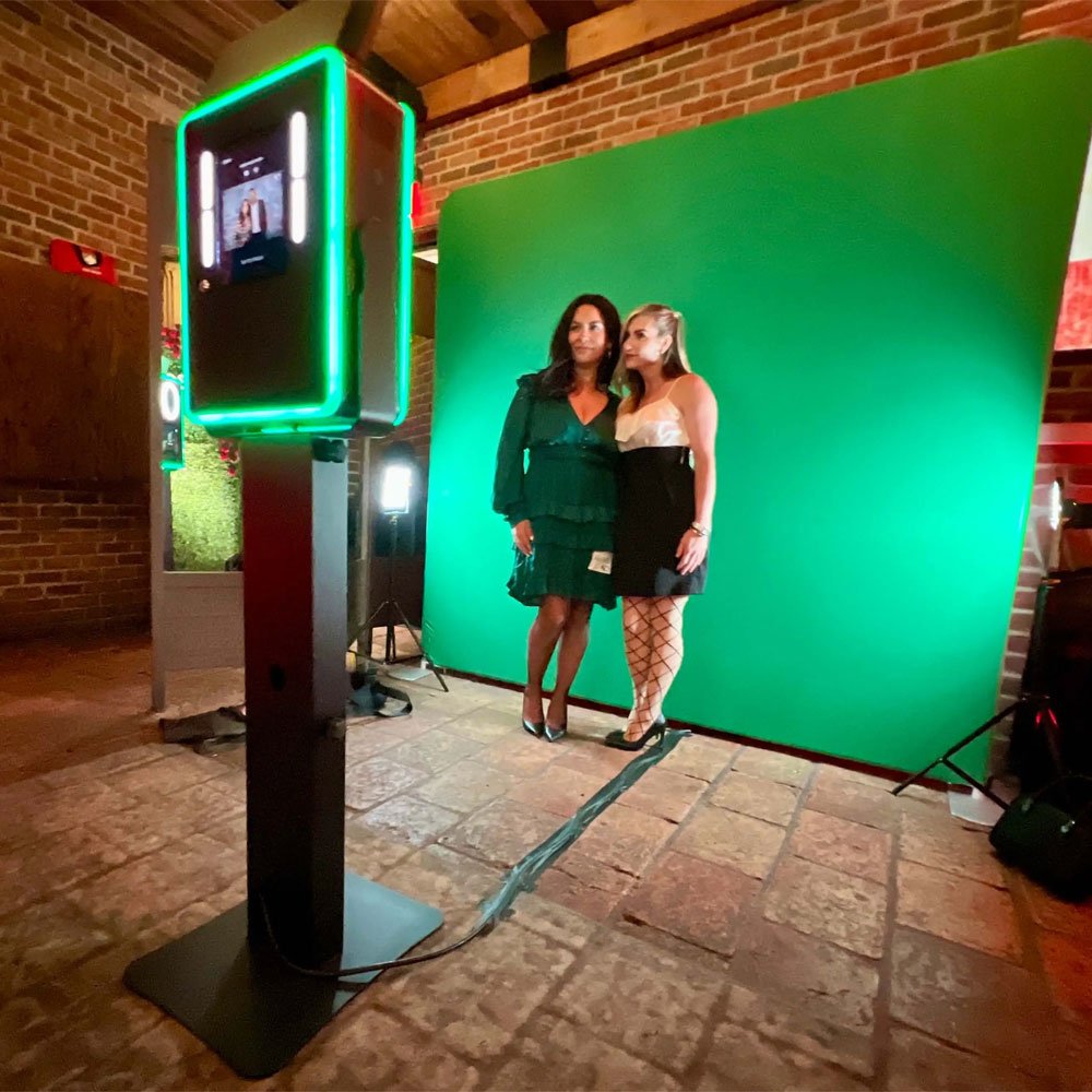 Two women posing for a photo in front of a green screen with a photo booth setup in an indoor space with brick walls.