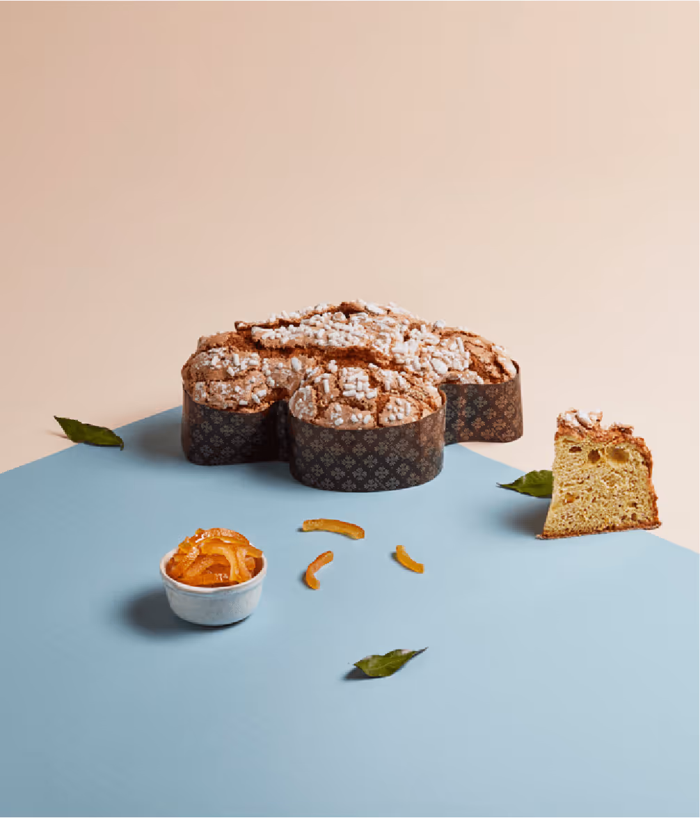 Colomba pasquale decorata con zucchero granulato, una fetta tagliata e scorze d'arancia candita in ciotola e sparse.