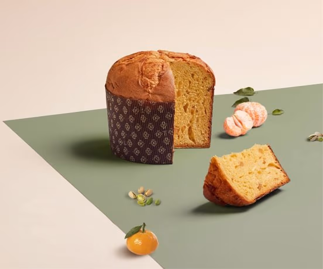 Panettone artigianale con fetta tagliata, accompagnato da pistacchi e mandarini su superficie verde e beige.