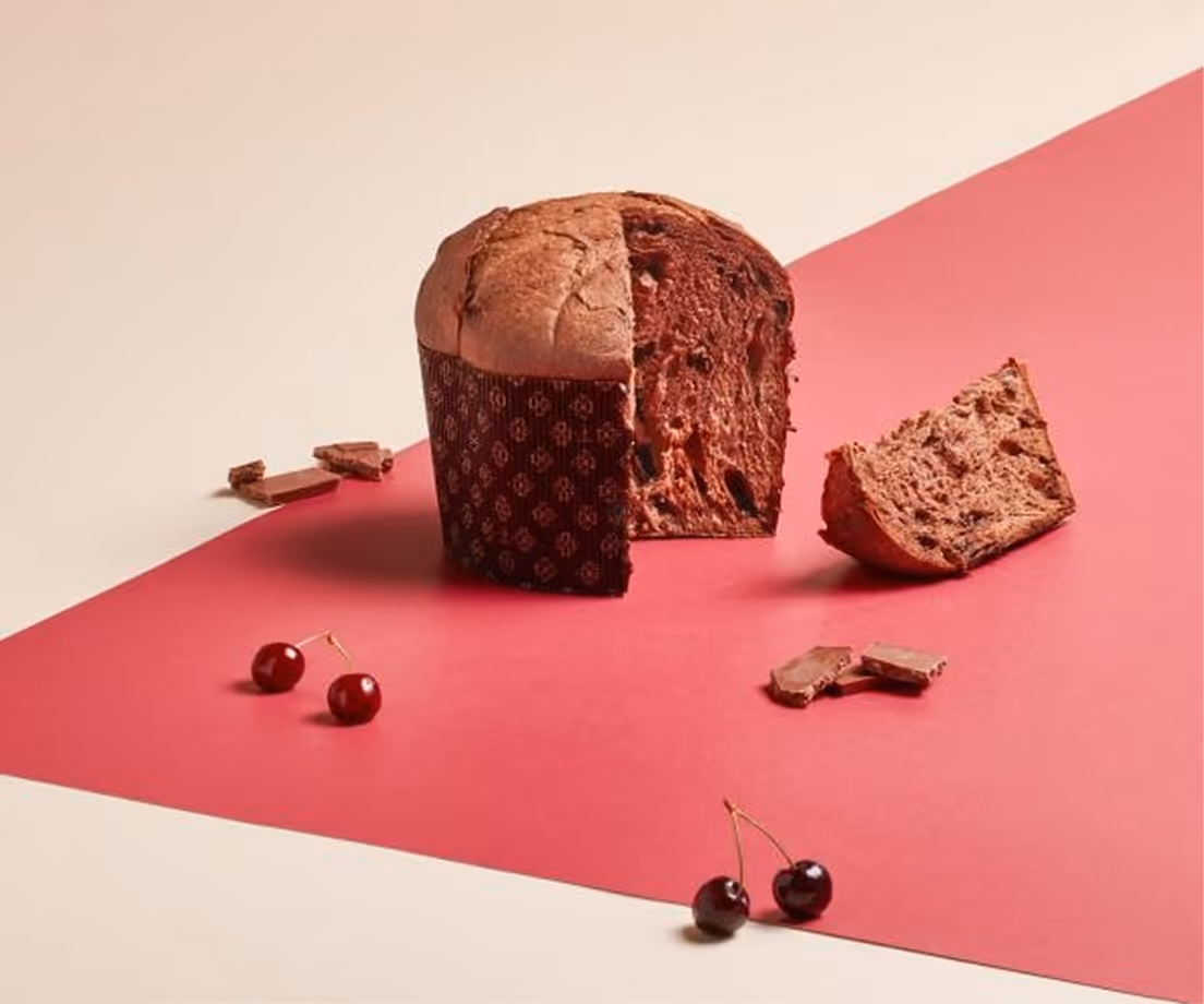 Panettone al cioccolato con amarene e pezzi di cioccolato su sfondo bicolore rosa e beige.