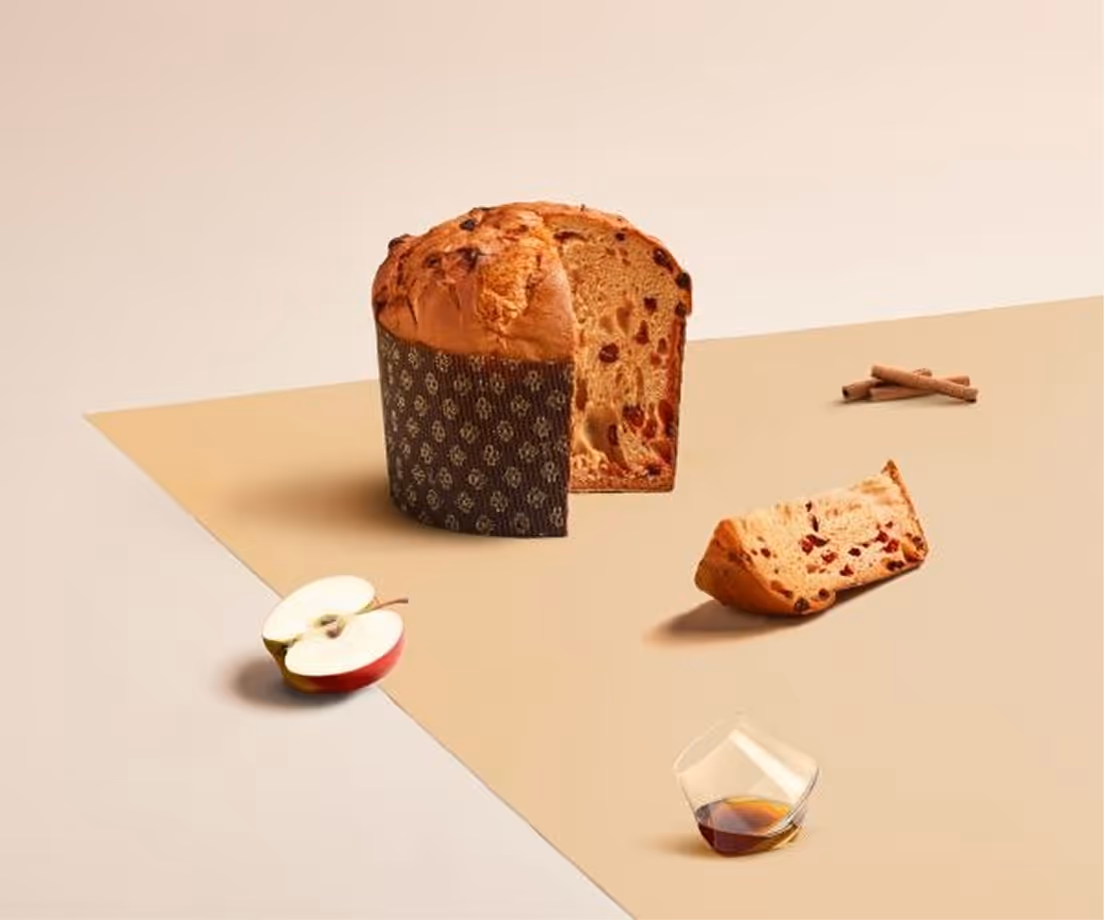 Panettone artigianale con frutta secca, una fetta tagliata, una mela tagliata a metà, cannella e un bicchiere inclinato con liquore.