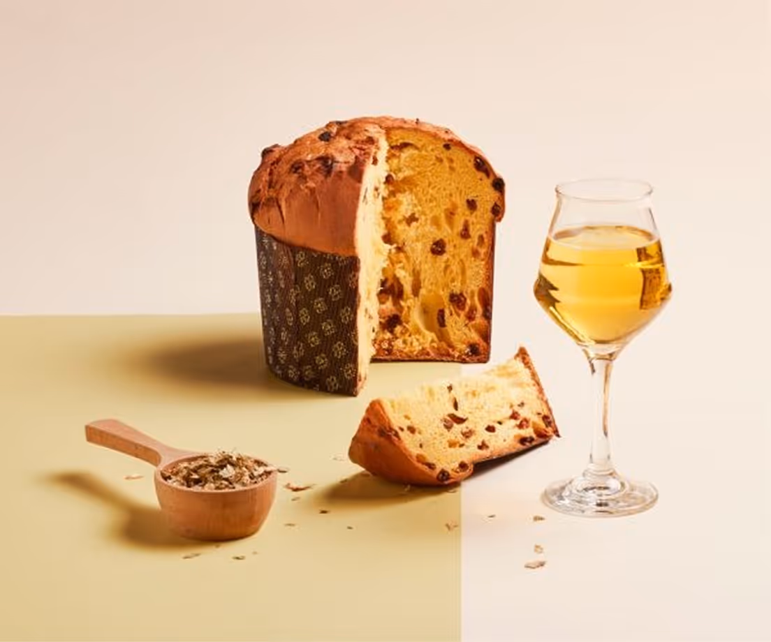 Panettone artigianale con uvetta e arancia candita, fetta tagliata, bicchiere di vino dorato e misurino di farina con lievito.