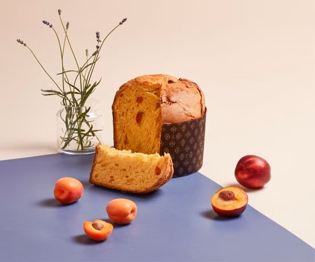 Panettone artigianale con pezzi di albicocca affettato, circondato da albicocche fresche e un vaso con rametti di lavanda.