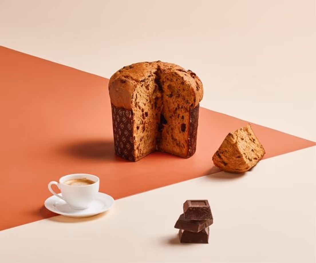 Panettone artigianale al cioccolato con tazza di caffè e pezzi di cioccolato su sfondo bicolore.