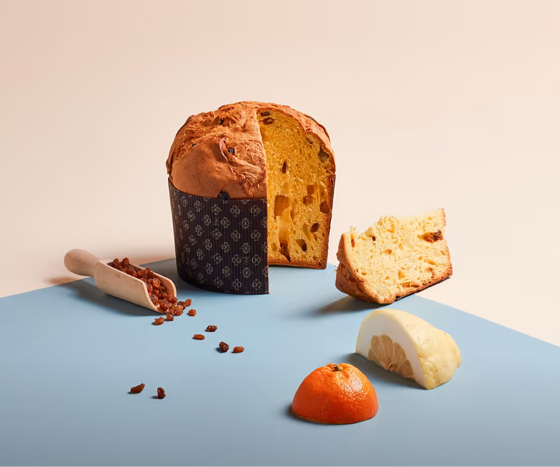 Panettone artigianale classico tagliato a fette con uvetta sparsa su una superficie blu, accanto a una fetta di arancia e una fetta di cedro.