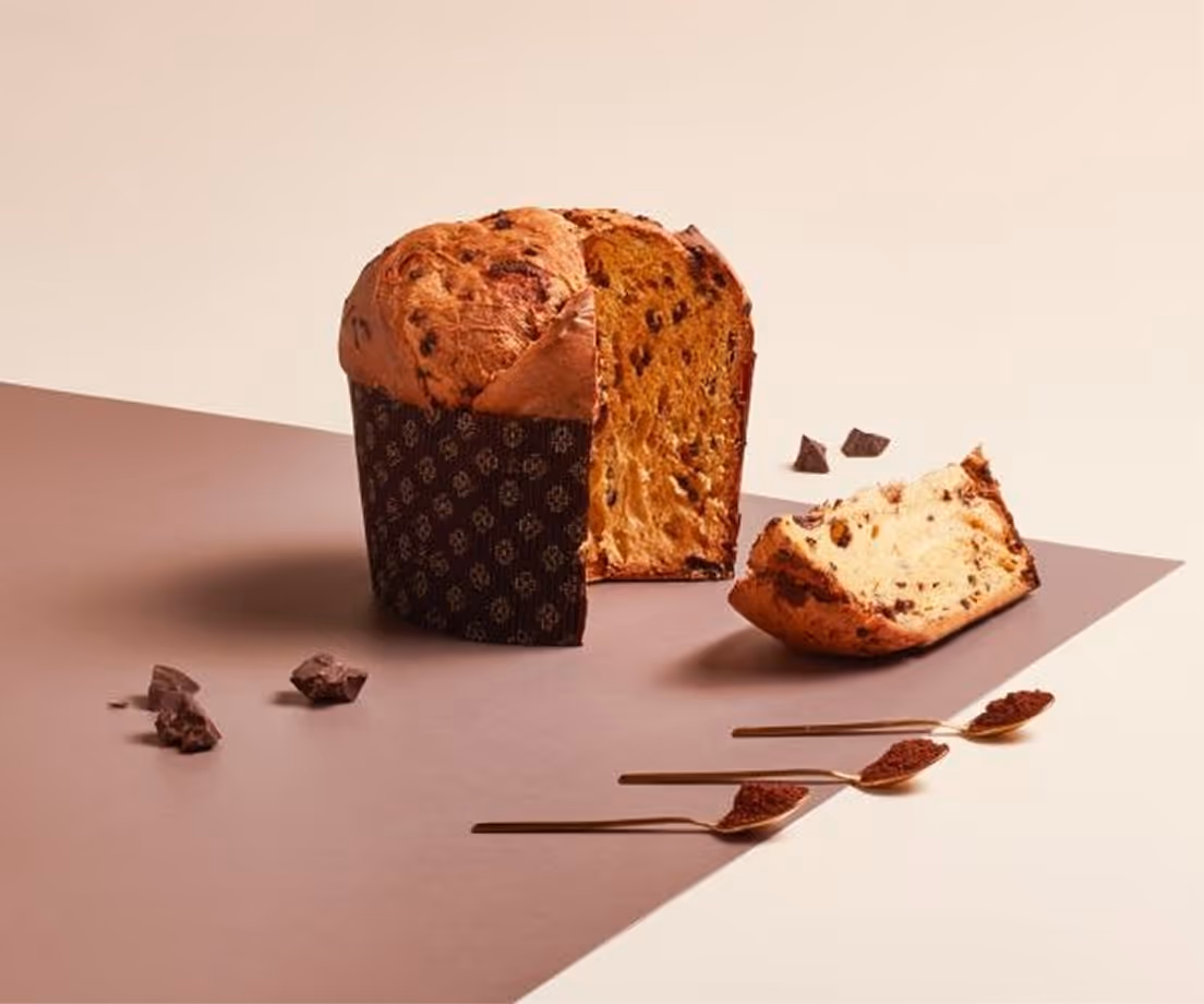 Panettone artigianale al cioccolato con una fetta tagliata e pezzi di cioccolato accanto su una superficie bicolore.