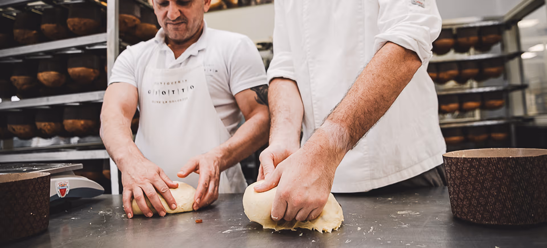 Due pasticceri impastano la pasta per panettone in laboratorio con teglie di carta sul tavolo.
