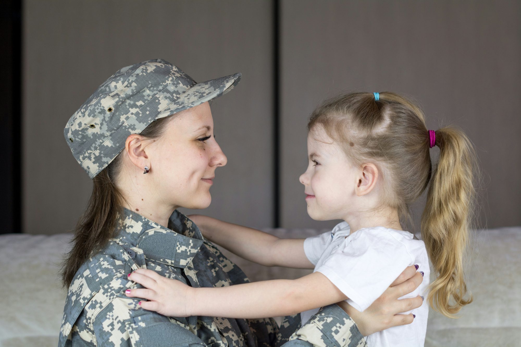 A mother in a military uniform and cap smiling face to face with her young daughter at home