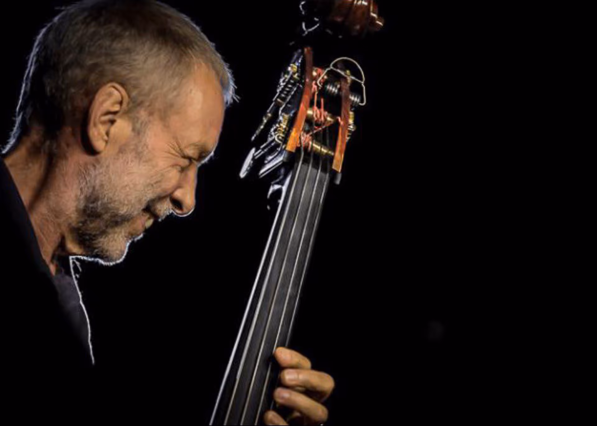 An elderly man plays a concentrated double bass against a black background, with a phiton resonator attached.