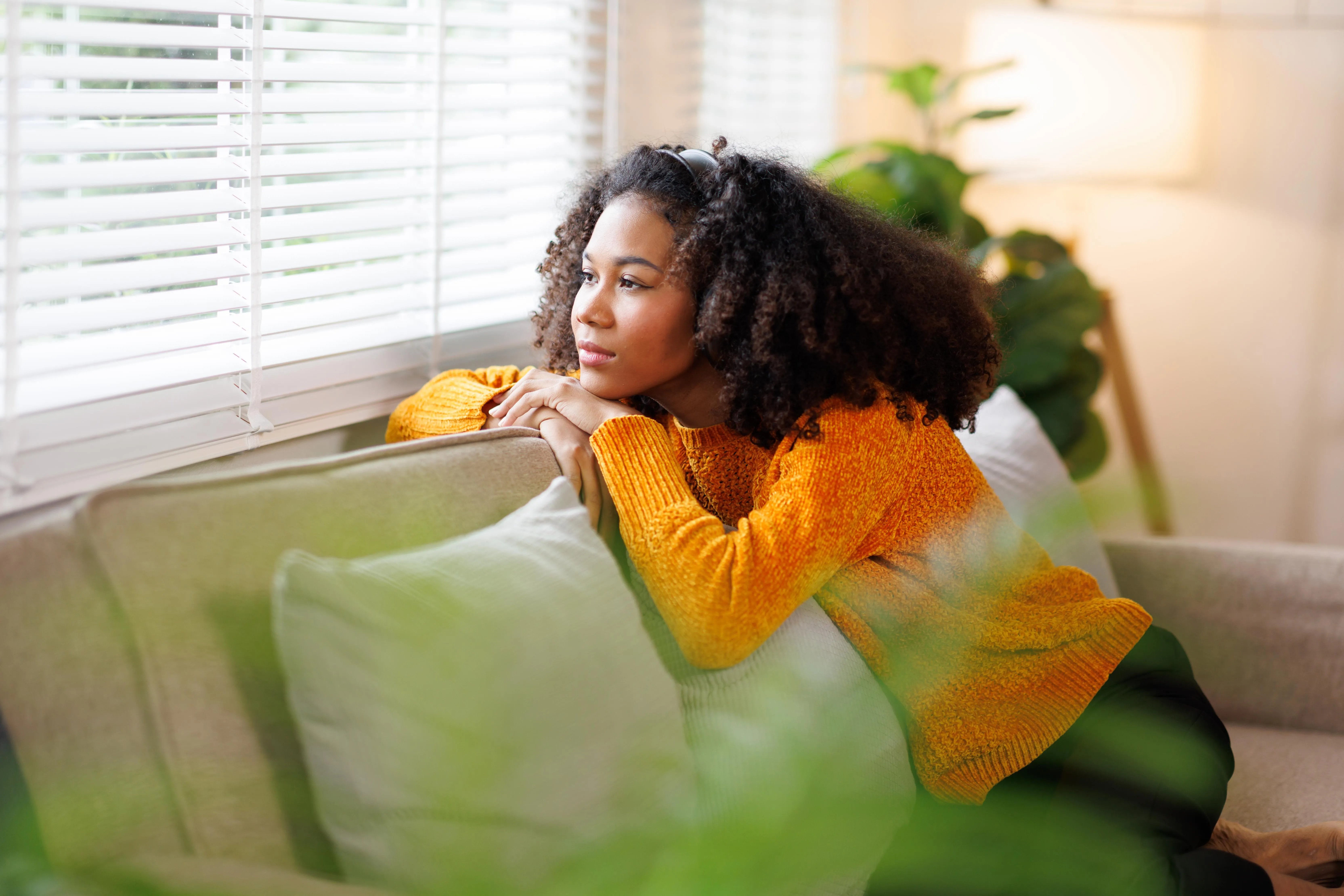 Young woman with curly hair looking thoughtfully out a window from a sofa, depicting a serene interior setting at Liberty at Estrella Falls in Goodyear, AZ.