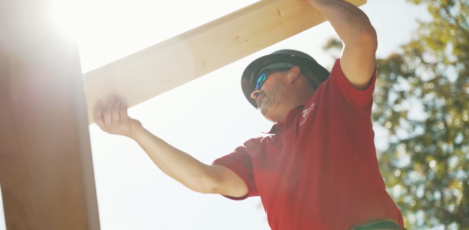 Worker in red shirt and safety helmet lifting wooden beam in sunlight