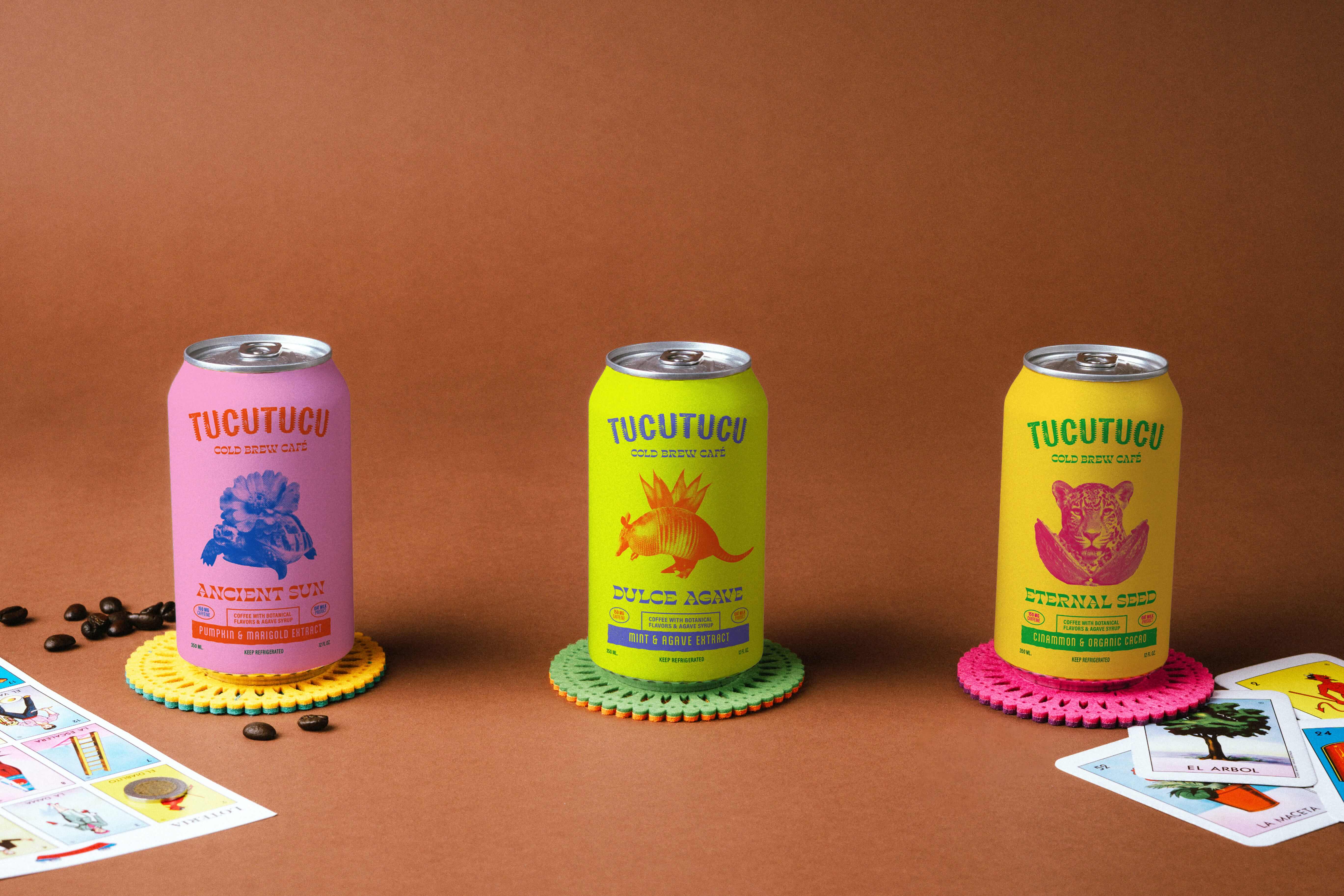 Three colorful Tucutucu cold brew café cans on crocheted coasters, surrounded by coffee beans and Lotería cards on brown background.