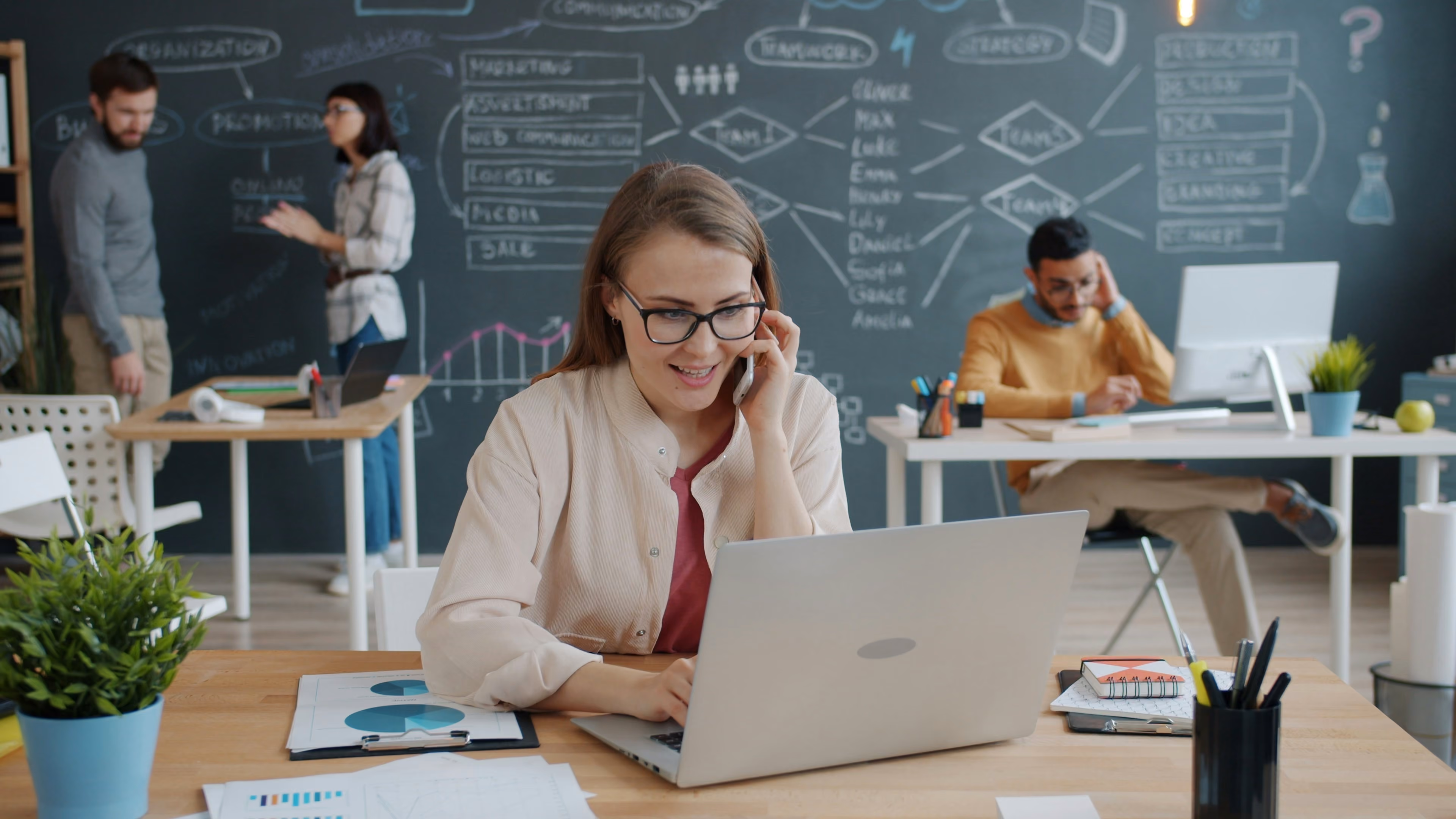 Woman with glasses talking on phone while using laptop in office with coworkers and a chalkboard wall.