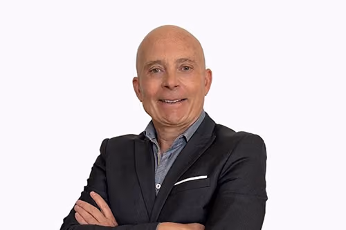 Smiling bald man in a black suit jacket with a blue shirt, arms crossed against a white background.