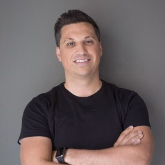 Man with short dark hair smiling, wearing a black t-shirt and watch, standing with arms crossed.