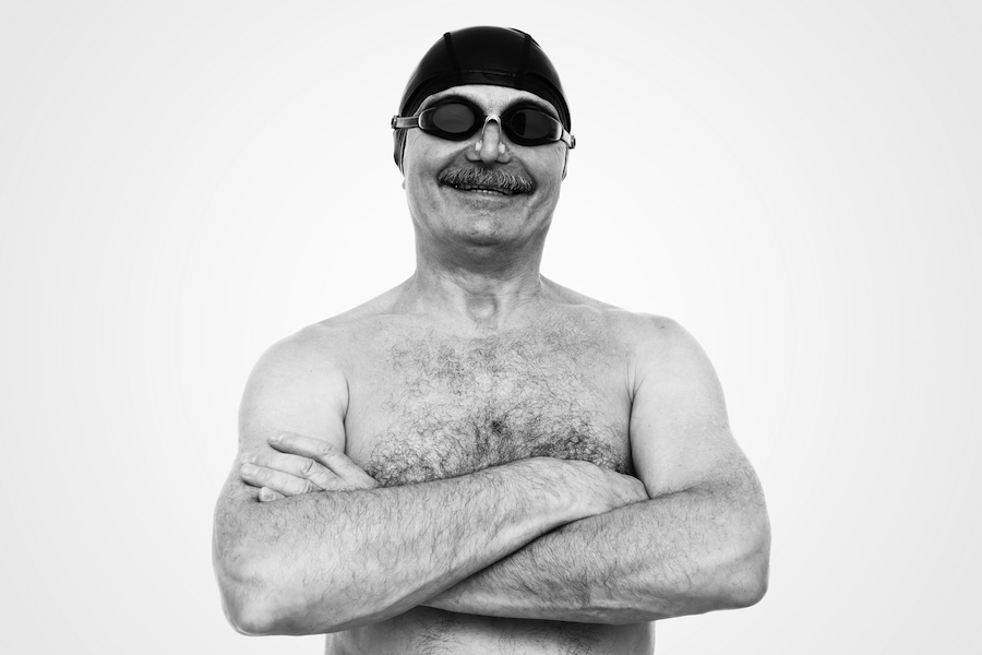 Man wearing a swim cap and goggles standing with arms crossed, black and white studio portrait