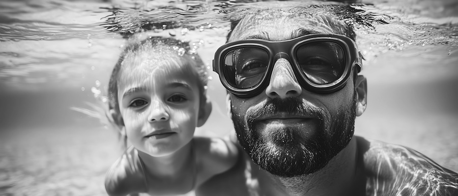 Bearded man in swim goggles and a young girl posing underwater in a pool, black and white