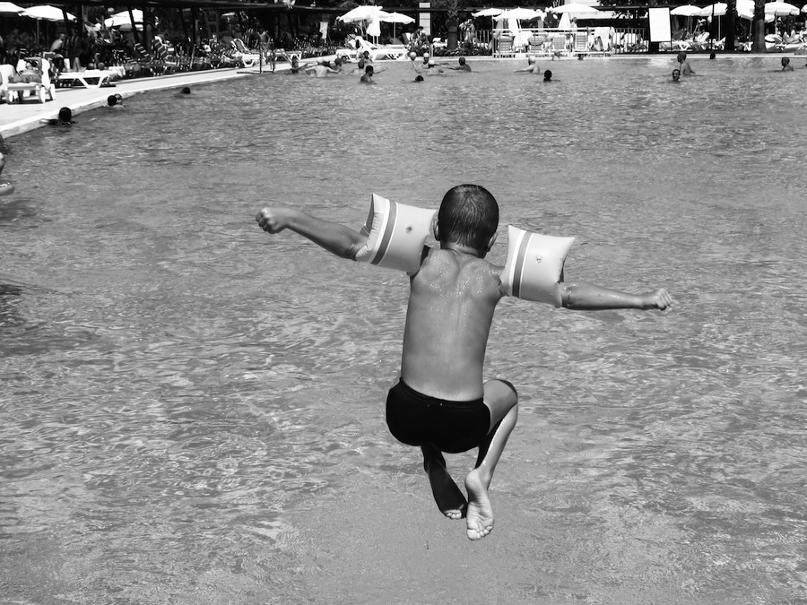 Child with arm floaties jumping into a crowded swimming pool from behind in black and white.