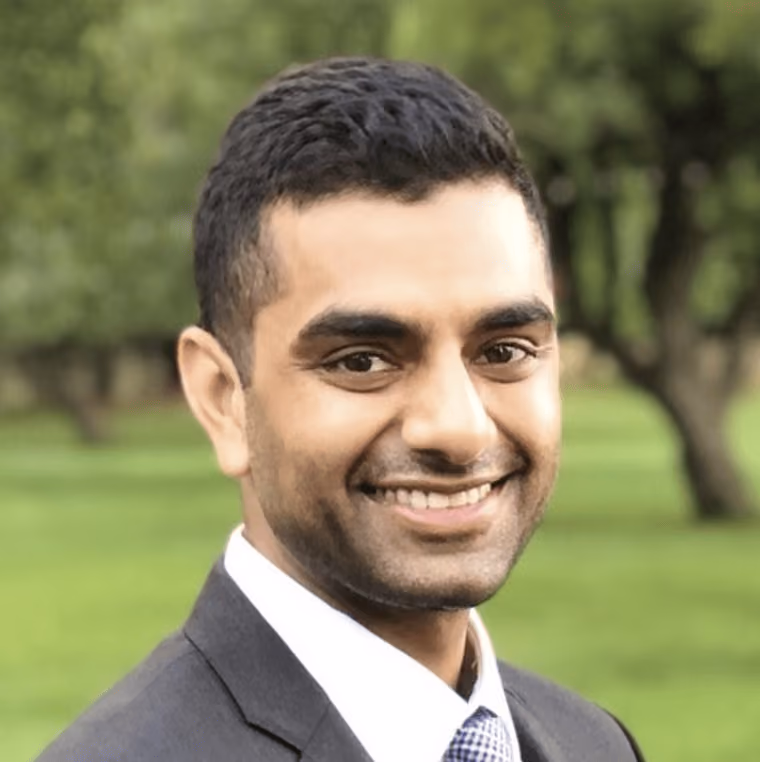 Smiling man wearing a suit and tie standing outdoors with greenery in the background.