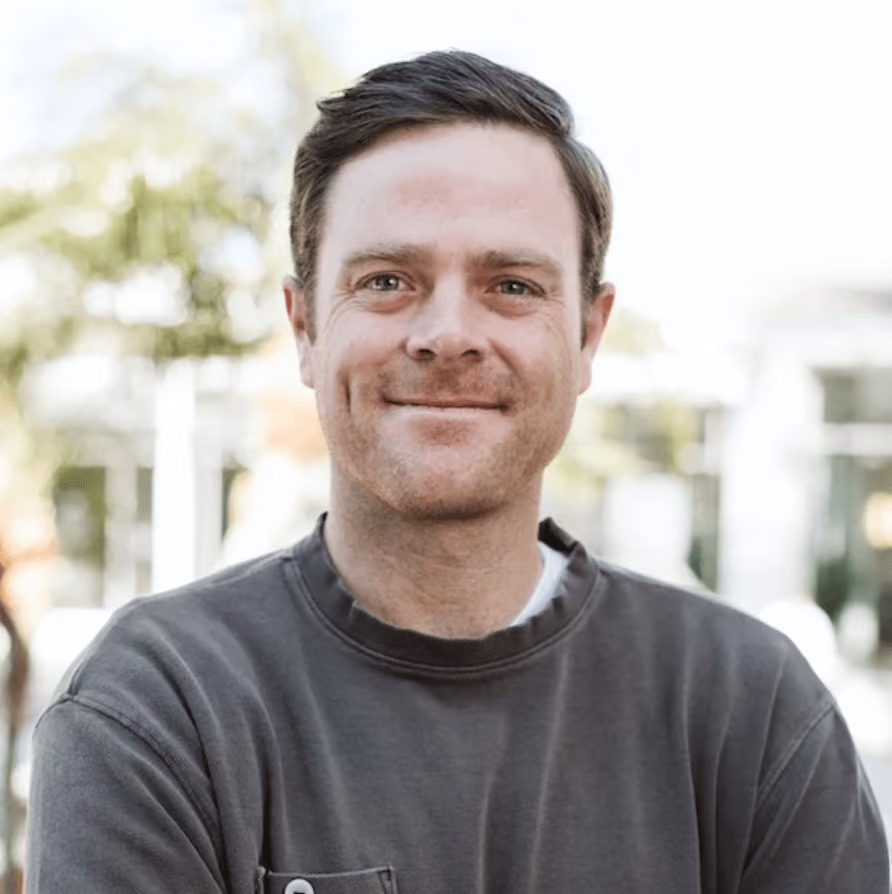 Smiling man with short brown hair wearing a dark gray sweatshirt, standing outdoors with blurred trees in the background.