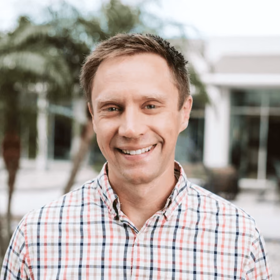 Smiling man with short light brown hair wearing a checkered shirt outdoors with blurred trees and buildings in the background.