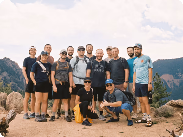 Group of thirteen hikers posing on a mountain trail with rocky terrain and forested hills in the background.