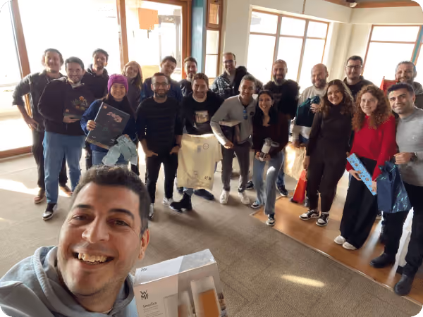Group of smiling people indoors, some holding gifts, with one person taking a selfie in the foreground.