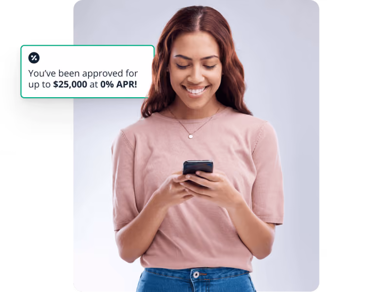 Smiling woman in pink shirt looking at her phone with a message showing approval for up to $25,000 at 0% APR.